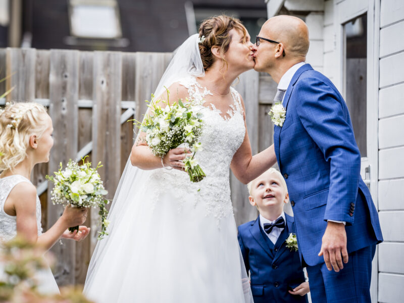 Bruidspaar kust elkaar terwijl bruidsjonker toekijkt. Fotograaf Karin Keesmaat van Kijk-Kunst fotografie