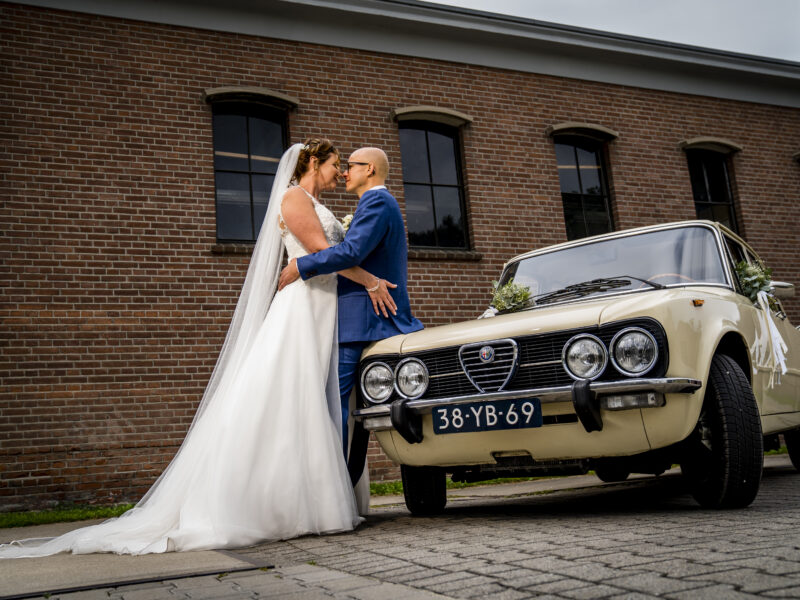 Bruidspaar pakt elkaar vast tegen een oude Alfa Romeo Giulia. Fotograaf Karin Keesmaat van Kijk-Kunst fotografie